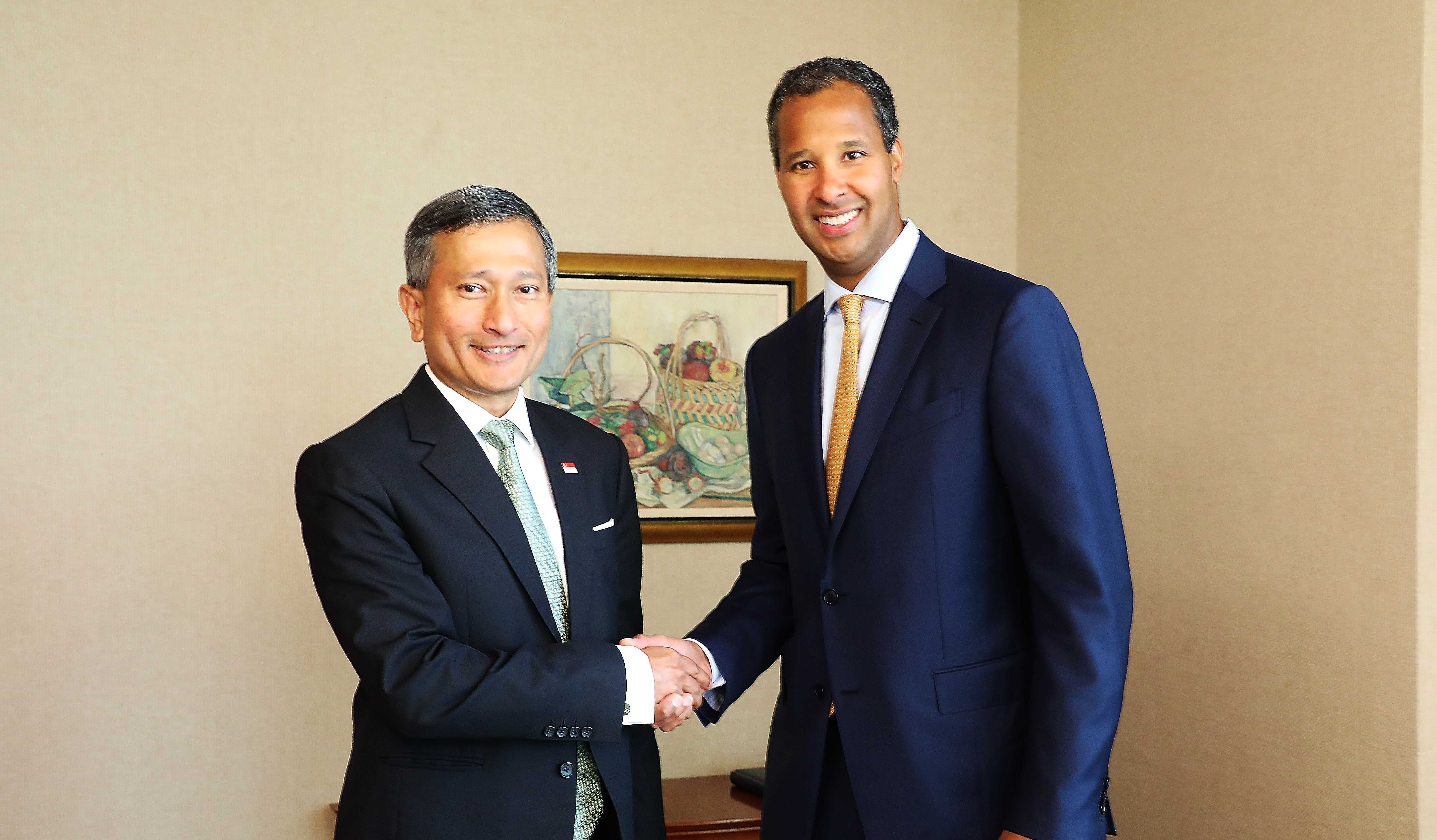 Two men in suits are shaking hands in front of a framed painting.
